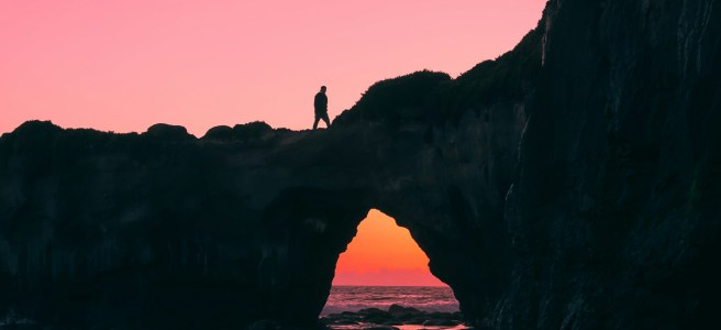 A person silhouetted against a sunset, walking over a natural stone arch bridge above the ocean, symbolizing the journey to close the execution gap and achieve personal growth.