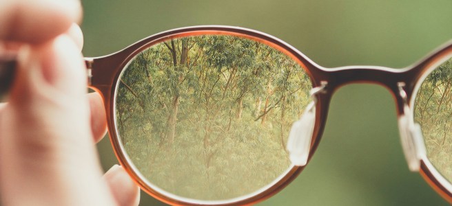 Hand holding glasses with a clear view of a green landscape through the lens, symbolizing focus, clarity, and personal growth.