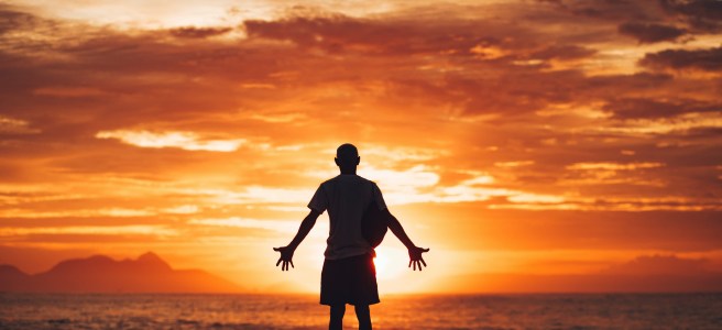Silhouette of a person standing on the beach at sunrise, reflecting on life, with arms open in front of the sun, symbolizing mindfulness and intentional living.