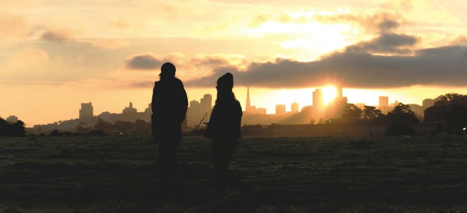 Silhouettes of two people walking in a field at sunrise, with a city skyline in the background, embodying the benefits of daily walking and mindfulness.