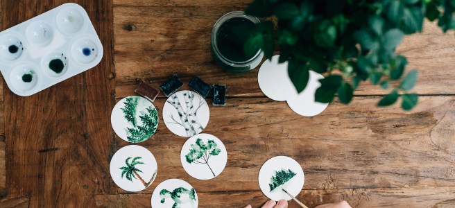 Person painting small watercolor trees on paper circles at a wooden table, surrounded by a watercolor palette and green plant.