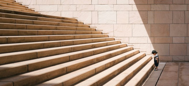 A small child stands at the bottom of a large staircase, symbolizing the challenge of taking the first step towards growth and overcoming obstacles.