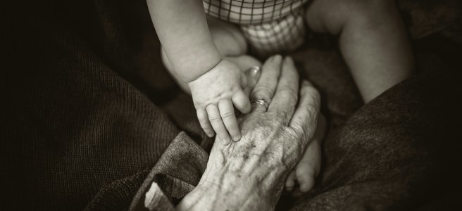 A black-and-white image of a baby’s hand touching an elderly hand, symbolizing generational wisdom and timeless growth across all ages.
